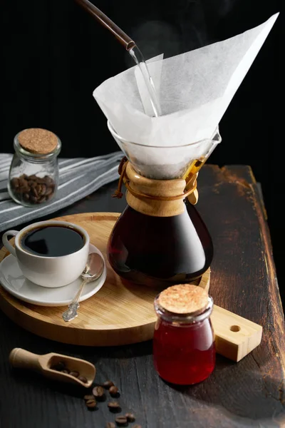 Häll över kaffebryggmetoden. Att hälla över kaffe med varmt vatten hälls från en vattenkokare. en kanna för att hälla över kaffe, en kopp kaffe på en ljus trä rund bricka, sylt i en burk, kaffe gr — Stockfoto