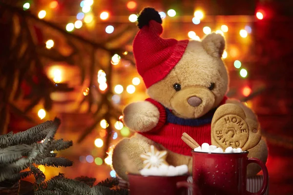 Composición navideña. Una taza roja con malvaviscos se encuentra frente a un oso de peluche con una gorra roja en el fondo de las luces de la guirnalda de Navidad. fondo de madera marrón — Foto de Stock