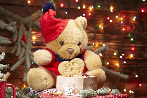 クリスマスの構図。テディベアは、クリスマスツリーの枝にあるクリスマスガーランドのライトを背景に、手作りのクッキーの箱を持って赤い帽子に身を包みます。茶色の木の背景 ストック画像