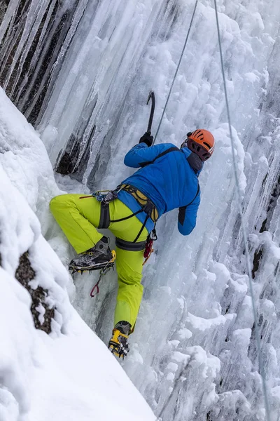 Ορειβάτης με μπλε μπουφάν, κίτρινο παντελόνι και κόκκινο κράνος σκαρφαλώνει στον παγωμένο καταρράκτη. Ο πάγος ανεβαίνει τον καταρράκτη. οπίσθια όψη. κατακόρυφη όψη — Φωτογραφία Αρχείου
