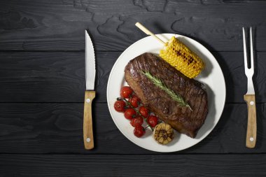 Mısır ve domatesle servis edilen bir tabakta ızgara biftek. Koyu arkaplan, üst görünüm, yatay fotoğraf