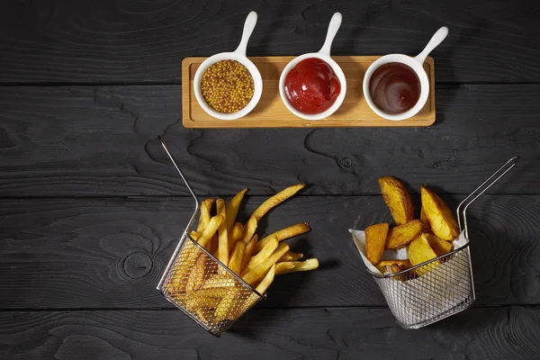 French fries and country potatoes in a basket and three different sauces: Ketchup, Mustard and Barbecue on dark background, top view, horizontal photo