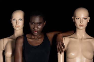 attractive young african american woman in bodysuit standing between mannequins and looking at camera on black  clipart