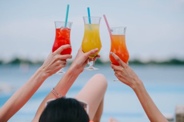 cropped shot of young female friends holding glasses with summer cocktails at beach clipart