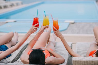 girls lying on chaise lounges and holding glasses with summer cocktails at poolside clipart