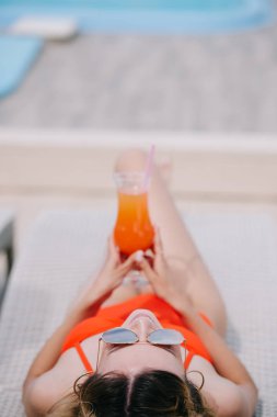 selective focus of girl in swimwear and sunglasses holding cocktail and resting on beach clipart