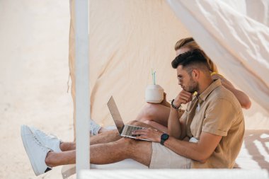 dizüstü bilgisayar kullanarak ve plaj bungalow kokteyl içme genç çift yan görünüm