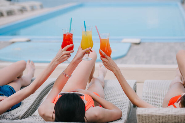 girls lying on chaise lounges and holding glasses with summer cocktails at poolside