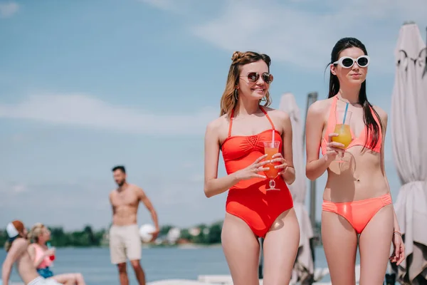 美丽的年轻妇女在太阳镜和泳装拿着杯子与鸡尾酒在海滩上 — 图库照片