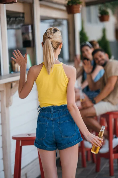 Visão Traseira Menina Segurando Garrafa Cerveja Acenando Mão Para Amigos — Fotografia de Stock