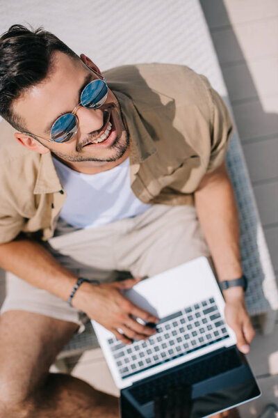 overhead view of young man in sunglasses using laptop and smiling at camera