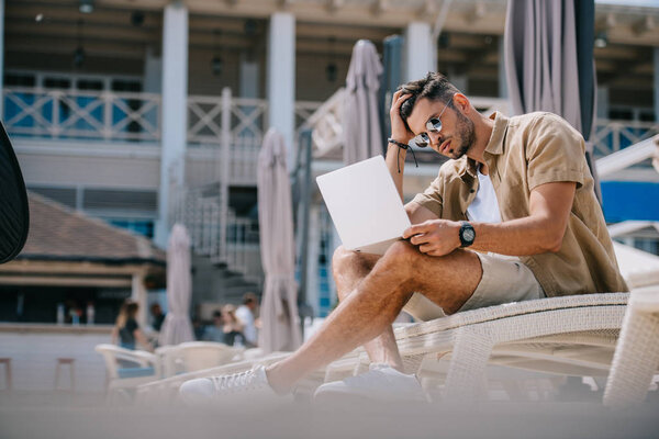 handsome young man in sunglasses using laptop while resting on chaise longue 