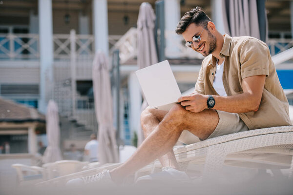 smiling young man in sunglasses using laptop while resting on chaise longue 