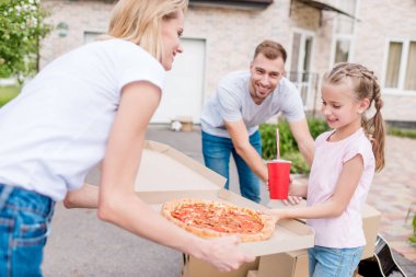 Anne holding kutusu pizza ile ve kızı alarak dilim pizza baba karton kutuları açma sırasında gülümseyen