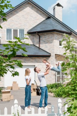side view of father raising up daughter while mother standing near in front of their new house  clipart