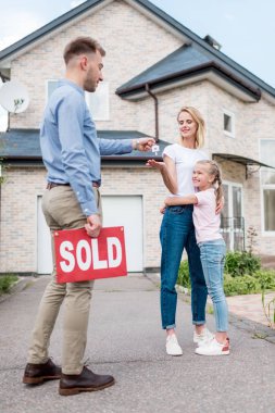 real estate agent with sold sign giving key to young woman with daughter in front of new house   clipart