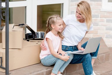 karton kutular ve yeni evin önünde gitar ile merdivenlerde dizüstü kullanarak kızı ile gülümseyen kadın 