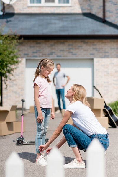 smiling woman tying shoelaces of daughter and man standing behind near cardboard boxes in yard of new cottage 