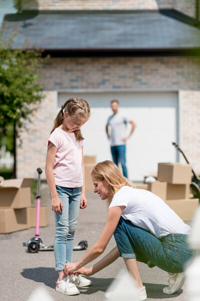 side view of smiling woman tying shoelaces of daughter and man standing behind near cardboard boxes in yard of new cottage 