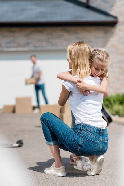 back view of woman hugging smiling daughter and man unpacking cardboard boxes behind in yard of new house 