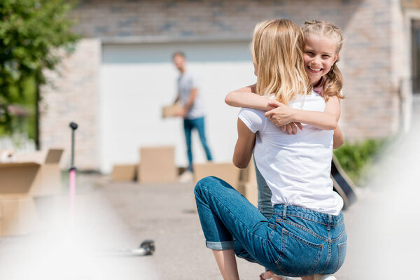 rear view of woman hugging smiling daughter and man unpacking cardboard boxes behind in yard of new house 