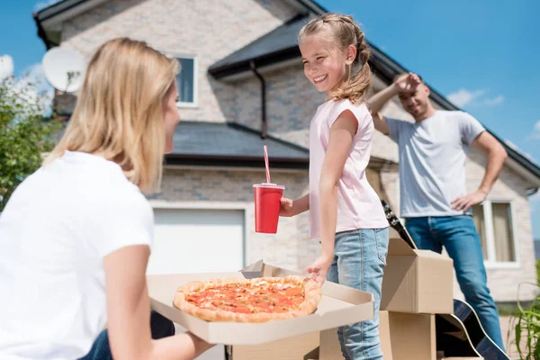 side view of happy mother holding box with pizza and daughter with cup of cola taking slice of pizza while father standing behind of new house