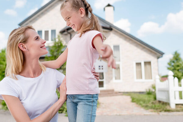close up view of key with trinket in hand of smiling little child embracing mother in front of new house 