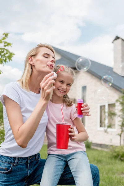 年轻妇女吹肥皂气泡 而她的女儿坐在附近的可乐杯 — 图库照片