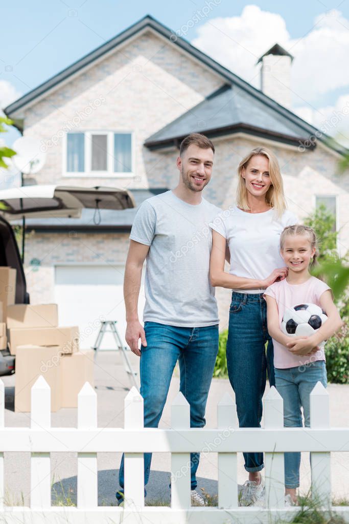 front view of happy family with little daughter holding soccer ball in front of new house 