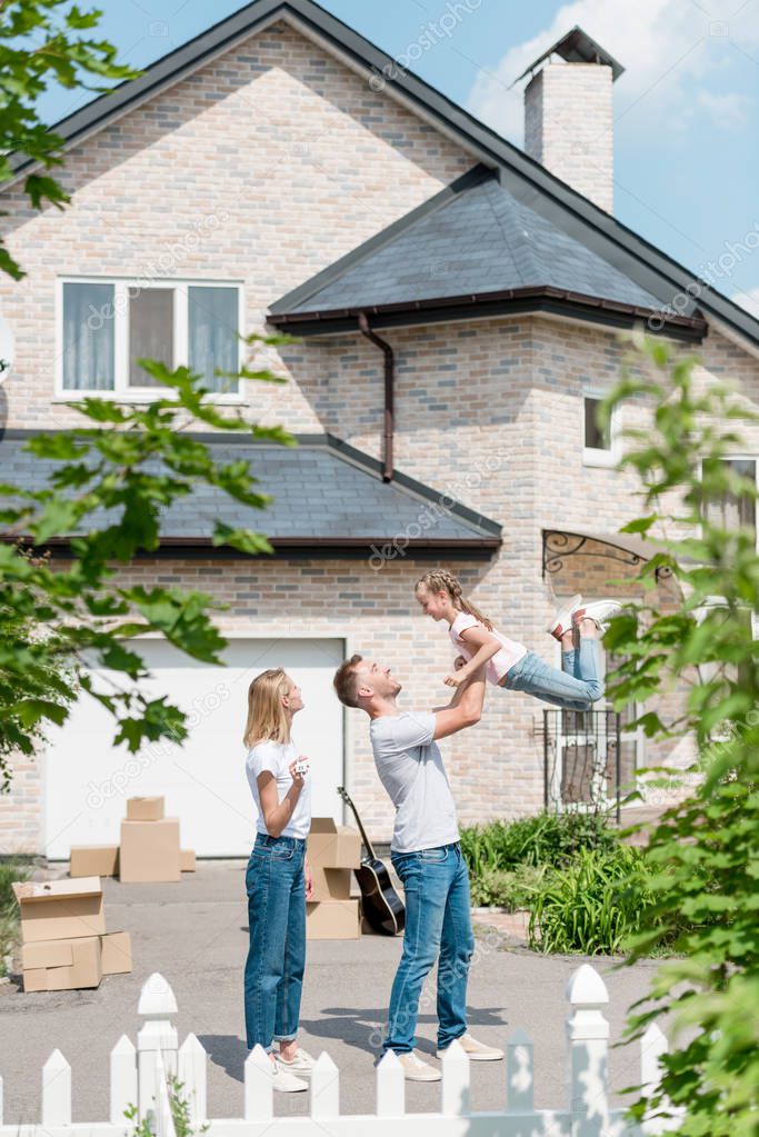 side view of father raising up daughter while mother standing near in front of their new house 
