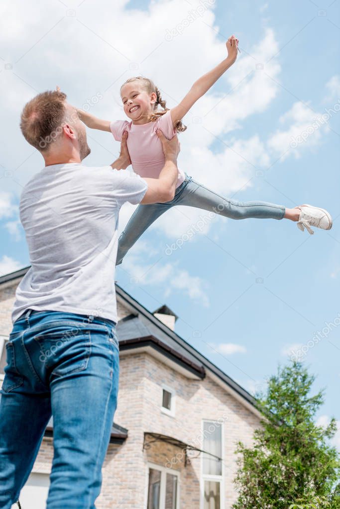 low angle view of father raising up smiling daughter with wide arms in front of house 