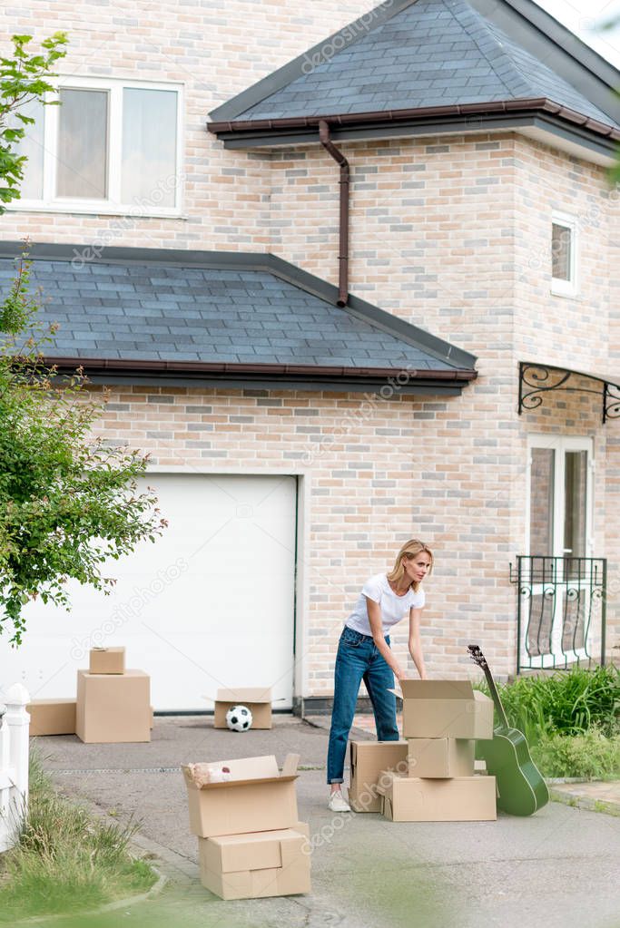 young woman unpacking cardboard boxes near guitar in front of new cottage 