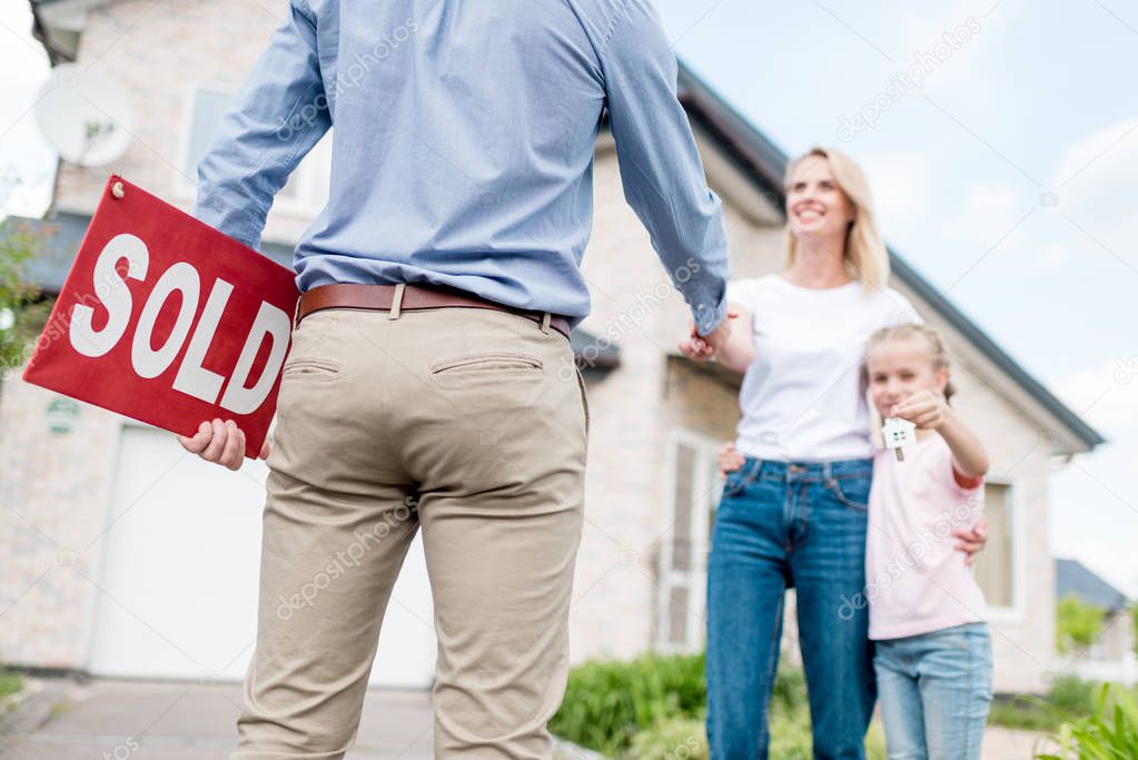 cropped shot of realtor with sold sign shaking hand of young woman with daughter in front of new house  