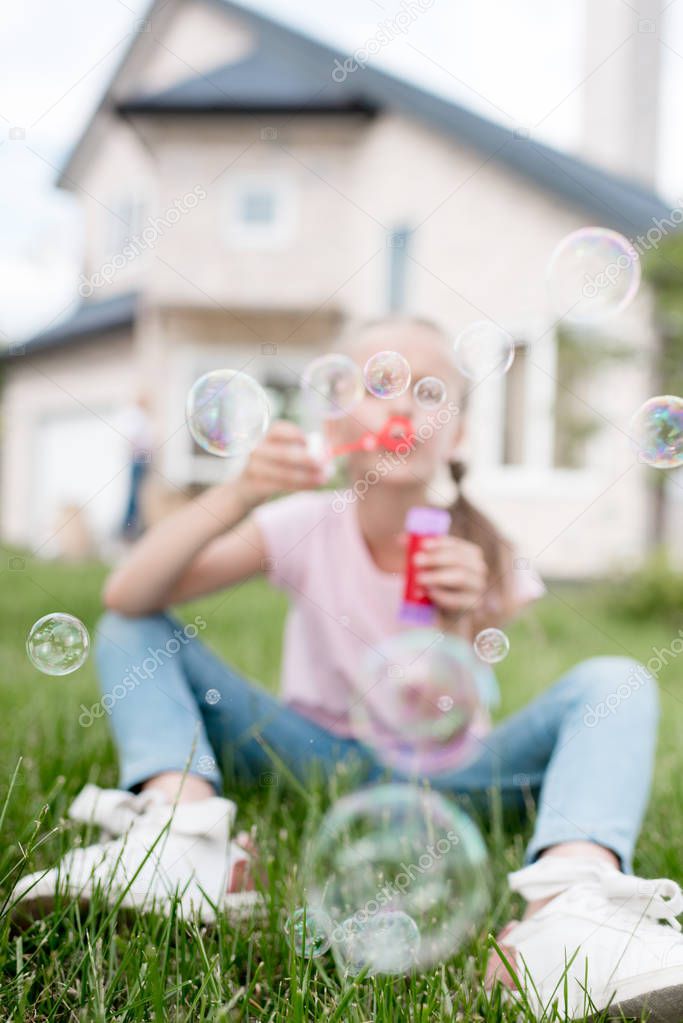 little child with soap bubbles sitting on lawn while her mother standing behind in front of house 