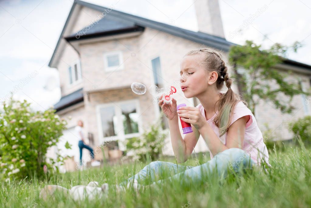 side view of little child blowing soap bubbles and sitting on lawn while her mother standing behind in front of house 