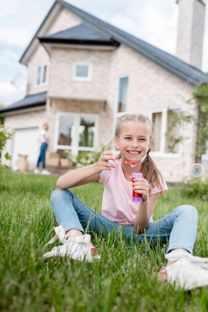 smiling little child with soap bubbles sitting on lawn while her mother standing behind in front of house 
