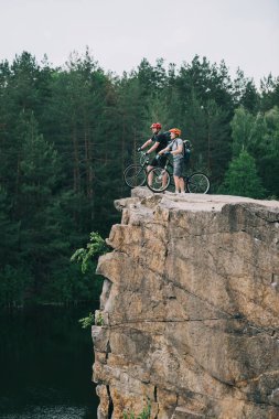 side view of young trial bikers standing on rocky cliff with blurred pine forest on background clipart