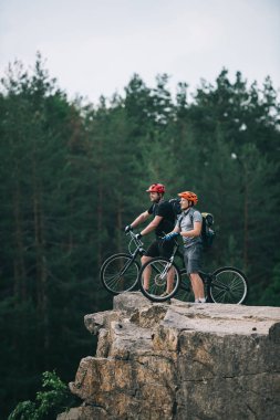 side view of happy young trial bikers standing on rocky cliff with blurred pine forest on background and looking away clipart