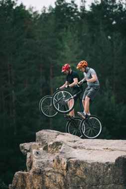 risky trial bikers standing on back wheels on rocky cliff with blurred pine forest on background clipart