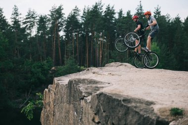 active young trial bikers standing on back wheels on rocky cliff with blurred pine forest on background clipart