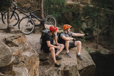 high angle view of young trial bikers relaxing on rocky cliff after ride and looking away clipart
