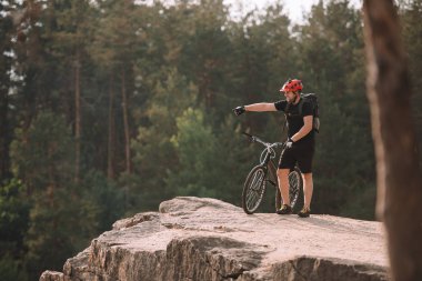 young trial biker standing on rocky cliff outdoors and pointing somewhere clipart