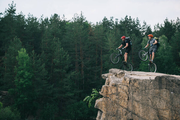 side view of young trial bikers with backpacks standing on back wheels on rocky cliff with blurred pine forest on background