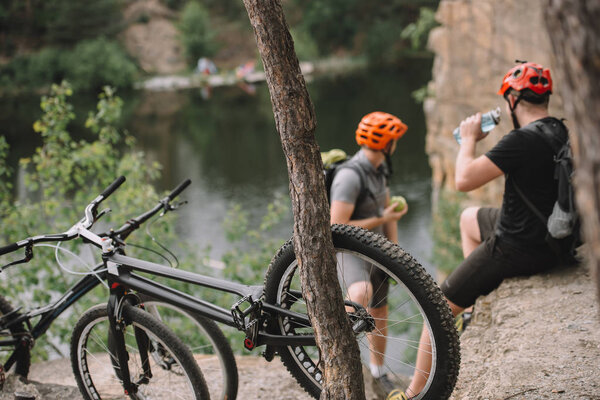 young trial bikers relaxing on rocky cliff after ride with bicycles on foreground