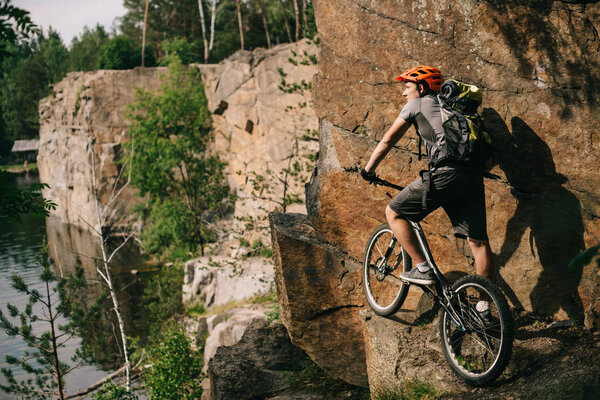 young trial biker with bicycle standing on cliff and looking at beautiful landscape