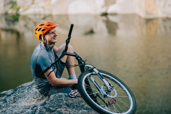 smiling young trial biker relaxing on rocky cliff over water