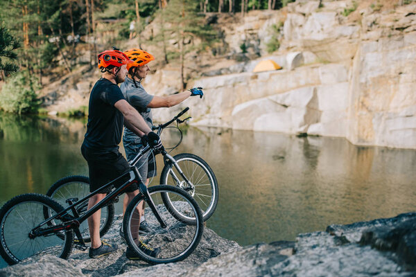 young trial bikers standing on rocky cliff in front of mountain lake and pointing at side