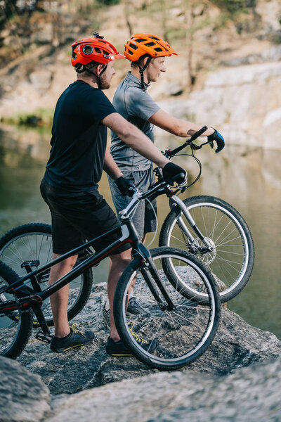 young trial bikers standing on rocky cliff in front of mountain lake and looking away