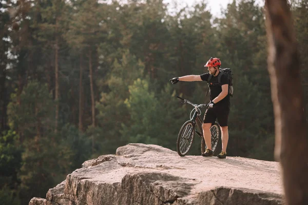 Young Trial Biker Standing Rocky Cliff Outdoors Pointing Somewhere — Stock Photo, Image