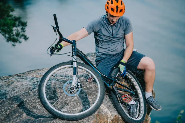 Young trial biker relaxing on rocky cliff over water with bicycle — Stock Image Young Trial Biker Relaxing Rocky Cliff Water Bicycle Stock Photo